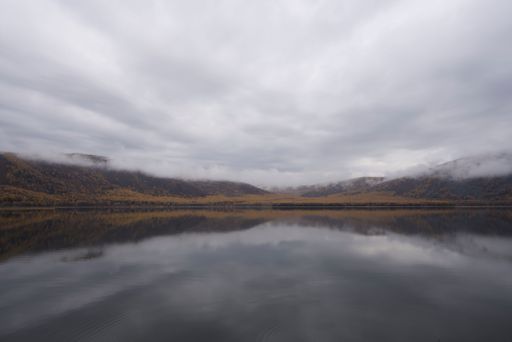 A serene landscape of a  lake surrounded by mountains with autumn foliage under a cloudy sky, in Nei Mongol, China