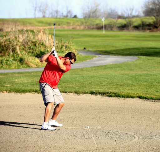 A man in his 30s playing golf, preparing to hit the ball from a sand bunker on a sunny day.