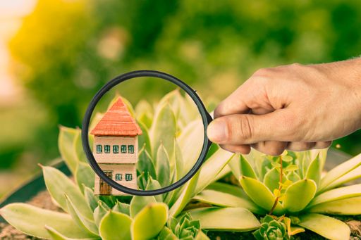 A hand with a magnifying glass and a wooden house model in green grass - selling real estate, investment concept