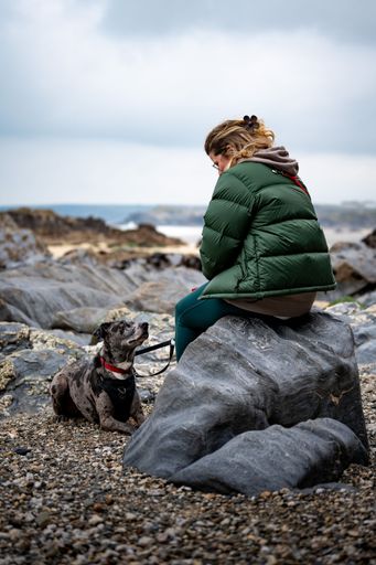 A woman sitting on a rock at a rocky beach with her dog on a leash looking at her, under a cloudy sky