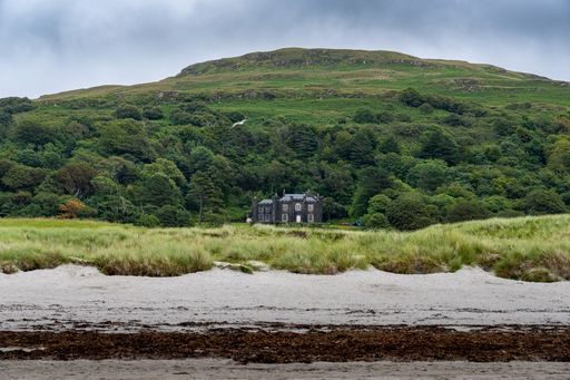 A scenic view of a mansion surrounded by lush greenery with a sandy beach in the foreground and a cloudy sky