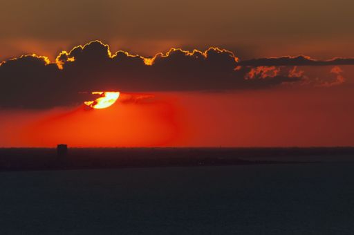 A stunning sunset over the ocean with the sun partially hidden by clouds and a distant cityscape.