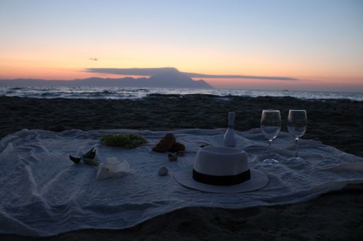 A romantic beach picnic setup at sunset with wine glasses, a hat, grapes, and a blanket