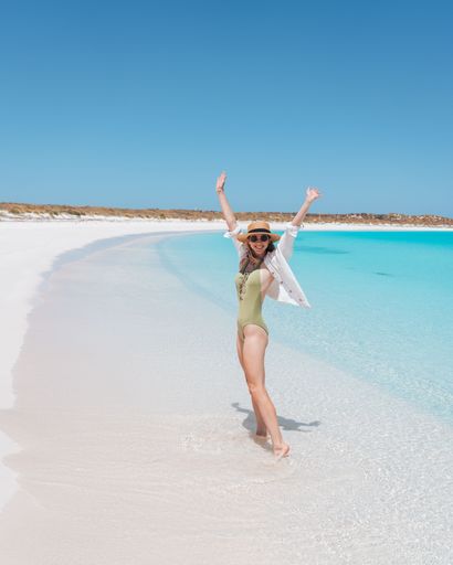 A woman in a swimsuit enjoying a sunny day on a pristine white sandy beach with turquoise water and clear blue sky.