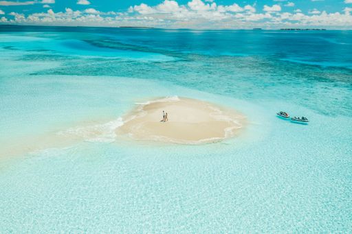 Aerial view of a couple standing on a small sandy island surrounded by turquoise water with boats nearby in the Maldives