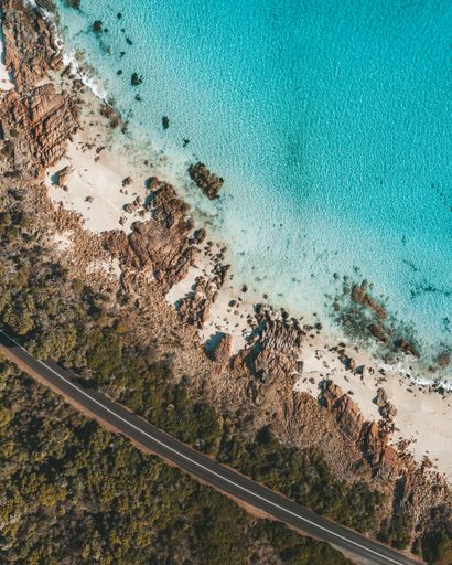 Aerial view of a coastal road alongside a turquoise blue ocean with rocky shore and lush greenery