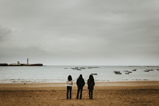 A wide shot of three people standing near the seashore with small boats floating in the sea