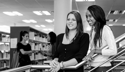 The two young women smiling and standing in a library with bookshelves in the background