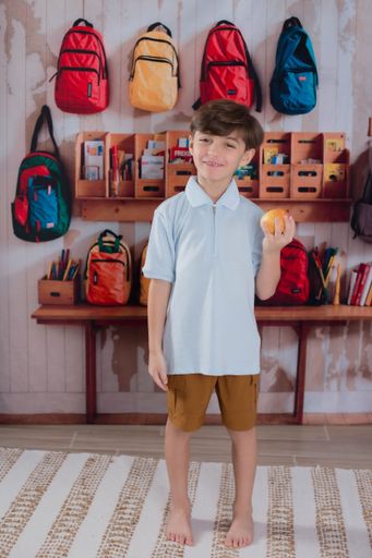 A young boy holding an orange stands in a classroom with colorful backpacks hanging on the wall.