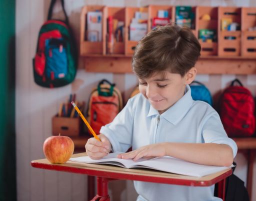 Young boy writing in a notebook at a classroom desk with an apple on the table and backpacks in the background