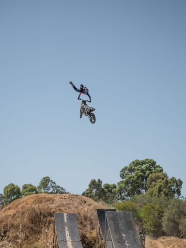 A motorcyclist performing an aerial stunt on a dirt bike with ramps and trees in the background under a clear blue sky.