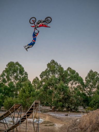 Motocross rider performing a mid-air stunt over a dirt ramp with trees in the background at sunset.