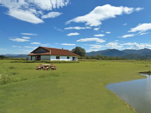 A scenic countryside view featuring a rustic house, picnic table, and a small pond with mountains in the background under a clear blue sky.