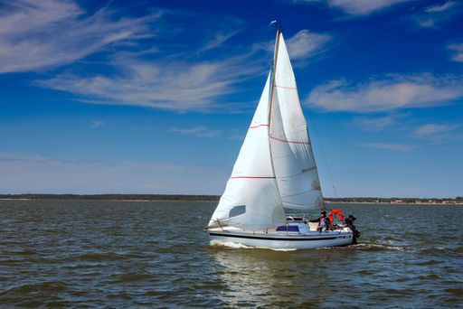 A sailboat with people sailing on a calm sea under a clear blue sky with scattered clouds