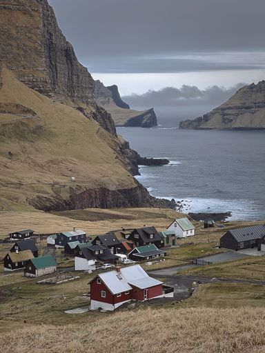 A scenic view of a small coastal village surrounded by mountains and the ocean under a cloudy sky in the Faroe Islands.