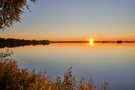 A serene sunset over a calm lake with a clear sky and lush vegetation in the foreground.