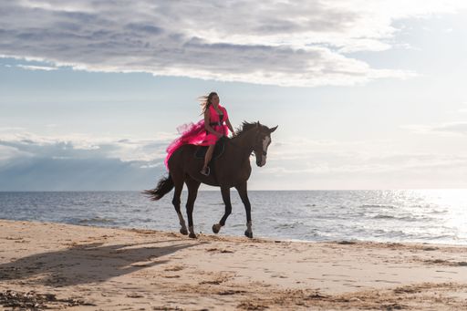 A woman riding a horse on the beach with a vibrant pink dress under a cloudy sky at sunset