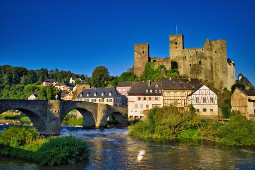 A scenic view of a historic castle and traditional houses along a river with a stone bridge under a clear blue sky in Germany.