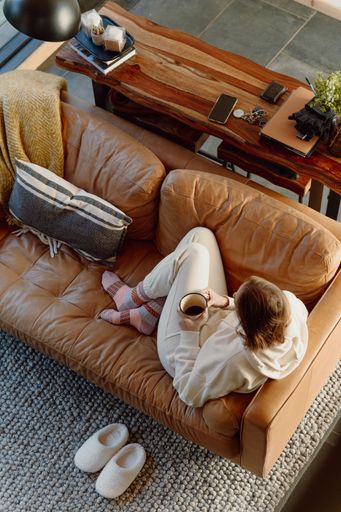 A vertical top view of a person relaxing on a leather couch holding a cup of coffee, wearing cozy socks and lounge-wear