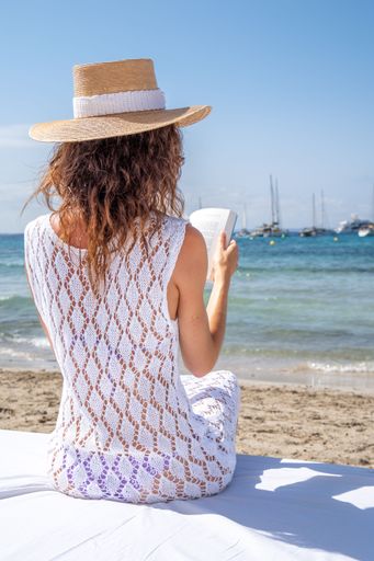 A woman in a straw hat reading a book on a sandy beach. Ibiza, Spain