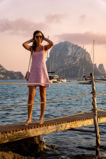 A woman in a pink dress standing on a wooden pier with sailboats in the background. Es Vedra, Ibiza
