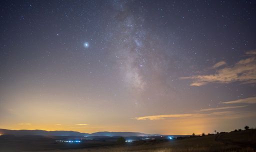 A stunning night sky with visible Milky Way over a rural landscape with distant mountains and scattered lights.
