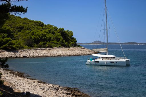 A sailboat anchored near a rocky shore and lush green trees under a clear blue sky on a sunny day