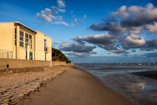 A serene beach scene with a building, sandy shore, and boats in the distance under a partly cloudy sky