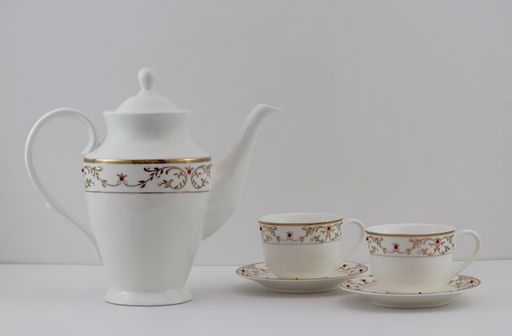 A closeup of a pair of ceramic cups and a teapot with intricate golden decorations on a white background