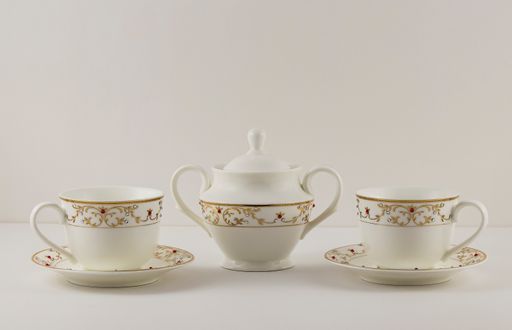 A closeup of a ceramic white teapot and two cups with intricate golden decorations on a plain background