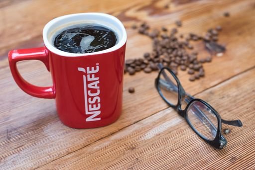 A selective focus shot of coffee in a Nescafe (Nestle) mug, glasses, and coffee beans on a table