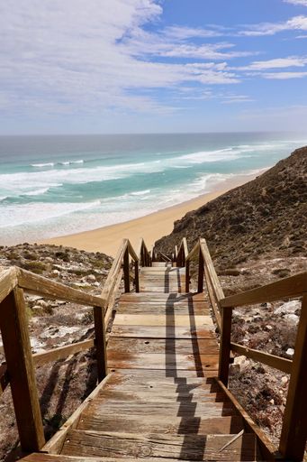 Wooden stairs leading down to a pristine beach with turquoise waves and a partly cloudy sky