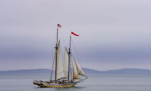 A sailing ship with American and red flag on calm waters under a cloudy sky, with distant hills in the background.