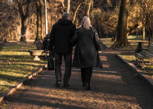 An elderly couple walking arm in arm down a park path during autumn with trees and benches around