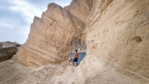 A young shirtless man standing on the edge of a cliff, looking through a hole in a rock formation at sea