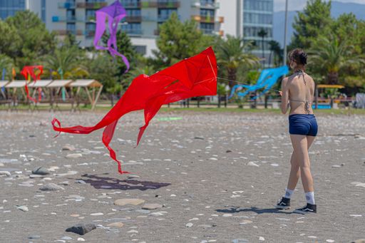 A young woman flying a red kite on a pebble beach with colorful kites in the background