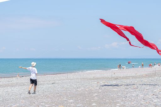 A young boy flying a red kite on a pebbly beach with a clear blue sky and ocean in the background