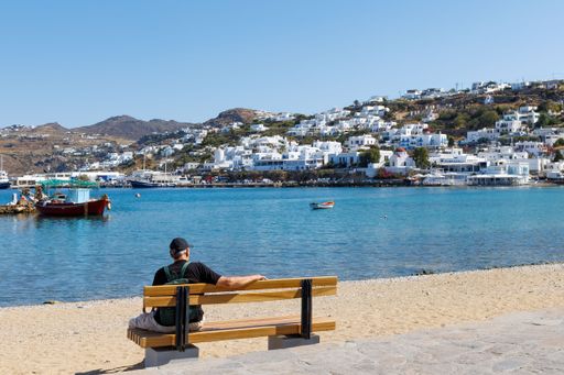 A man sitting on a bench by the seaside overlooking the town of Mykonos, Greece