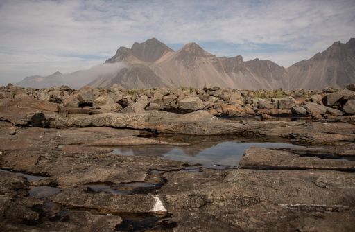 A rocky road with water patches leading to the mountains under a cloudy sky