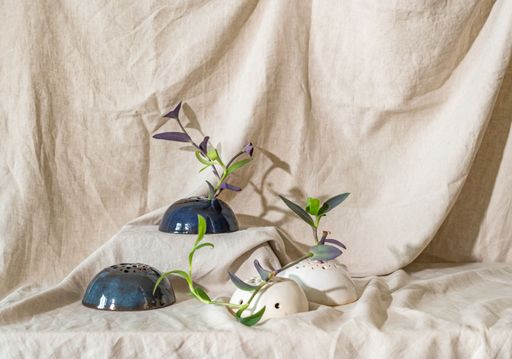 A minimalist still life of ceramic vases with green plants against a beige fabric backdrop.