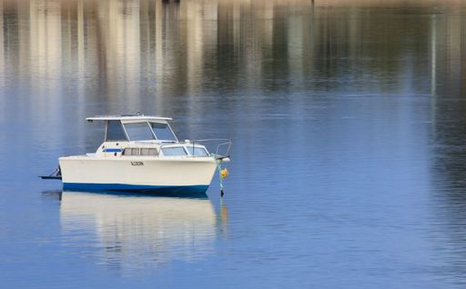 A small white and blue boat floating on calm water with a reflection of buildings in the background