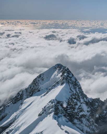 A breathtaking aerial view of a snow-capped mountain peak surrounded by clouds under a clear blue sky.