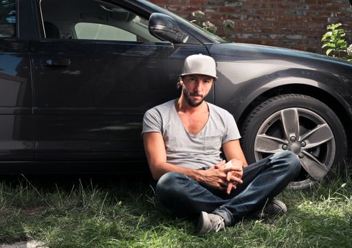 An adult Italian man wearing a cap and leaning on a black car parked in a field