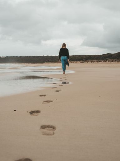 A woman walking barefoot on a sandy beach leaving footprints behind on a cloudy day.