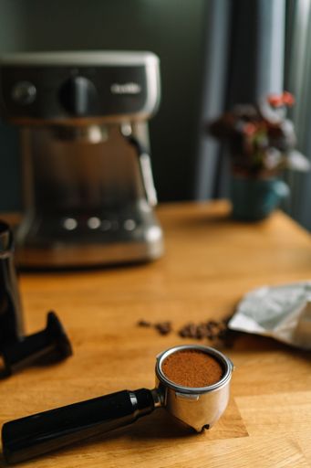 A close-up of a coffee portafilter filled with ground coffee on a wooden table with a blurred espresso machine in the background.