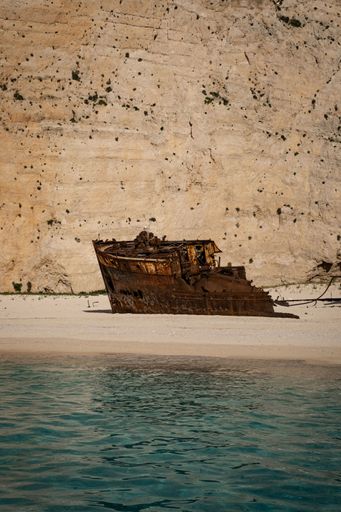 A Rusty shipwreck on the sandy shore of Navagio Beach in Zakynthos, Greece with clear turquoise water and a rocky cliff backdrop. Vertical shot.