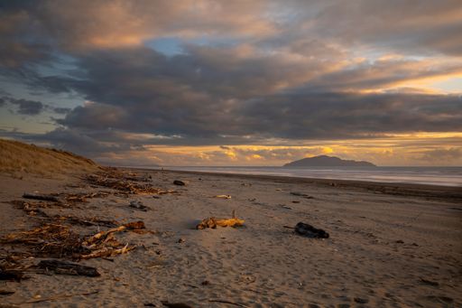 A dramatic sunset scene with driftwood scattered along the shore and Kapiti Island in a distance, in Otaki Beach on the Kapiti Coast of New Zealand