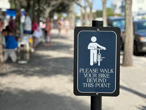 A Sign instructing cyclists to walk their bikes beyond a certain point on a busy sidewalk with blurred people and trees in the background.