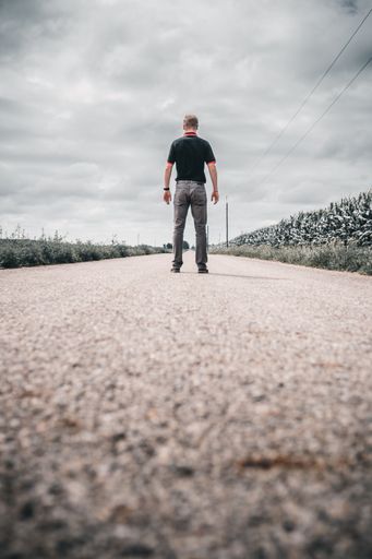 A man standing on an empty rural road under a cloudy sky, with fields on both sides.