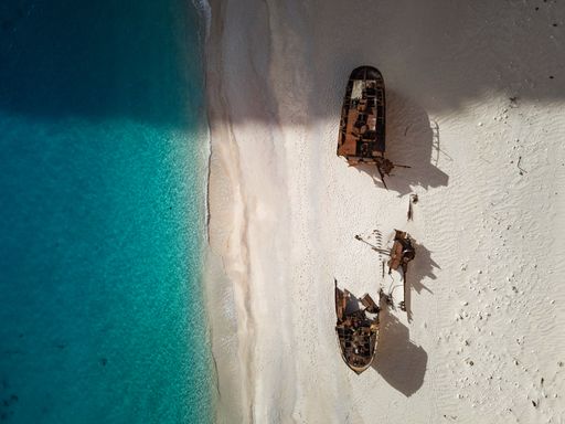 Aerial view of a shipwreck on a sandy beach with clear blue water, showcasing the contrast between the rusted ship and the pristine surroundings.